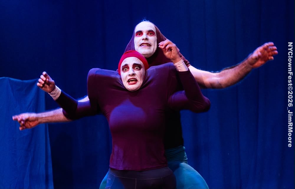 Two white-faced bouffon performers in maroon hoods, one stacked behind the other so their four arms reach out from one body.
